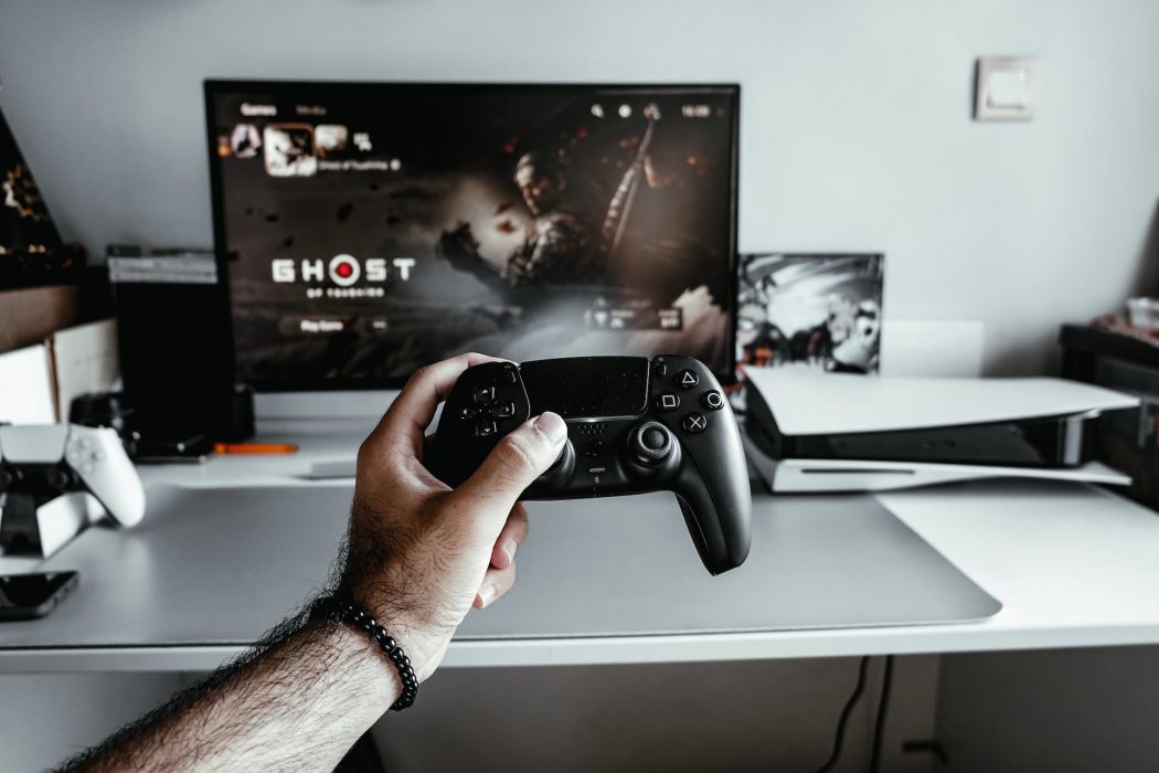 Man holds a ps5 controller in fron of the tv, a ps5 can also be seen on the right.